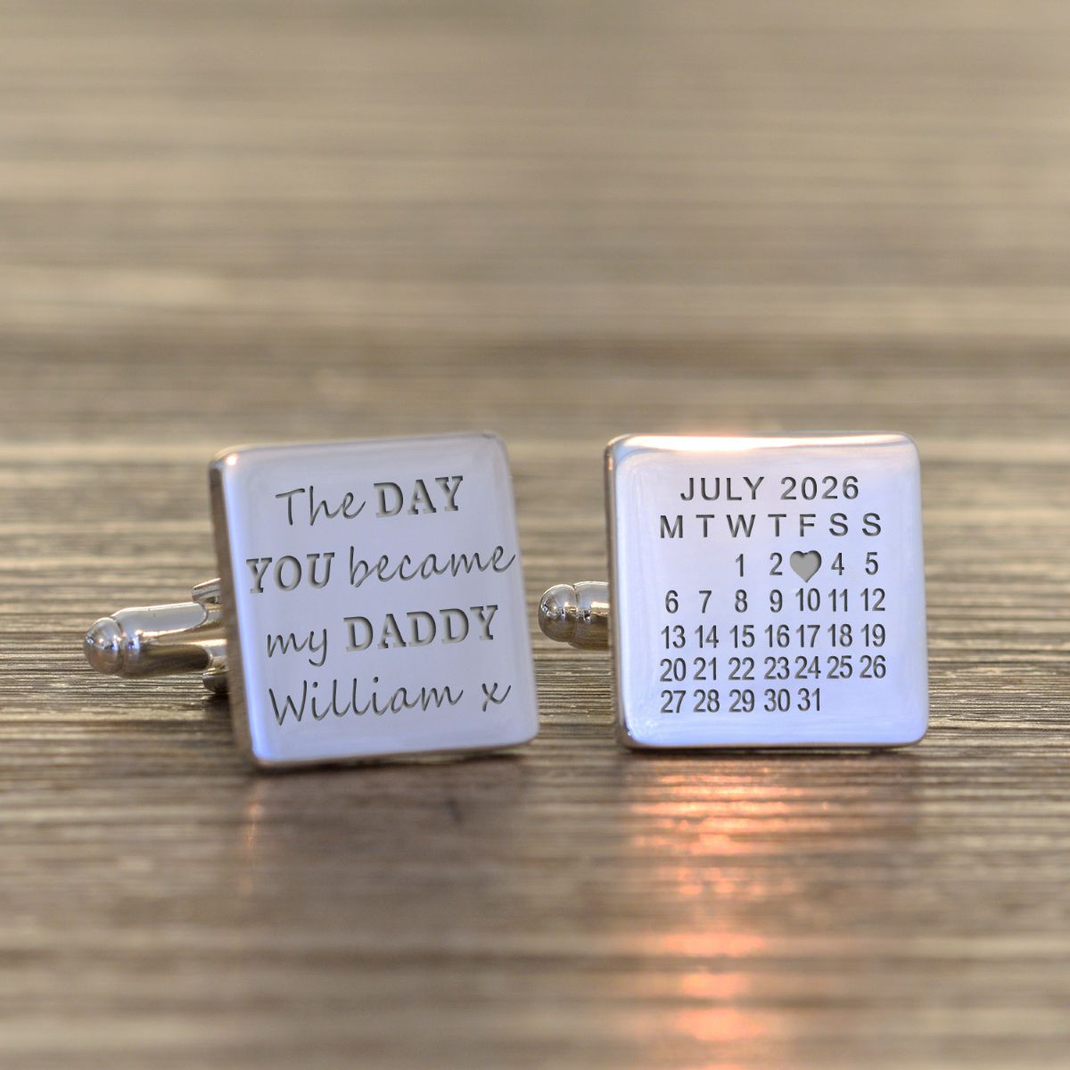 Two silver cufflinks with engraved text on a wooden surface, one says 'The DAY YOU became my DADDY William x' and the other a calendar with the 3rd July 2026 marked with a love heart.