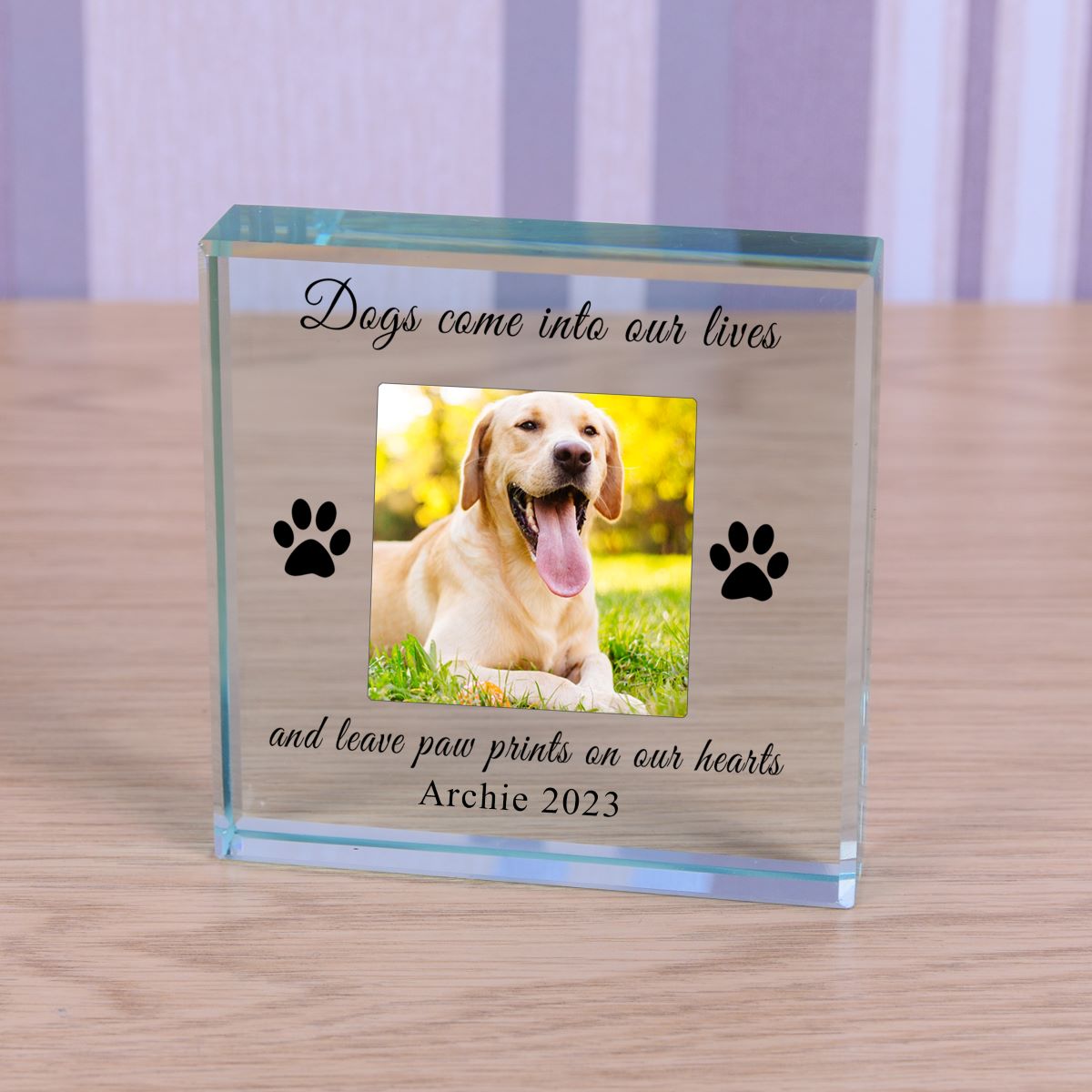 A personalised glass pet memorial block on a light wood tabletop. The clear block features a photo and a laser-engraved black text reading 'Dogs come into our lives and leave paw prints on our hearts Archie 2023', flanked by two black paw prints.