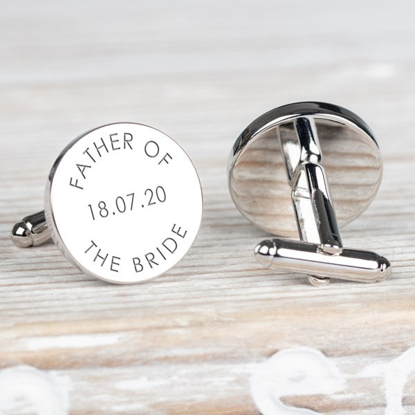 A pair of round silver cufflinks on a light wood surface. One cufflink is shown from the front with engraved text reading 'FATHER OF THE BRIDE' and a date.