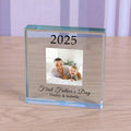 A personalised clear glass square block on a light wood tabletop, featuring engraved text '2025', a custom photo of a father and child, and 'First Father's Day Daddy & Isabella'.