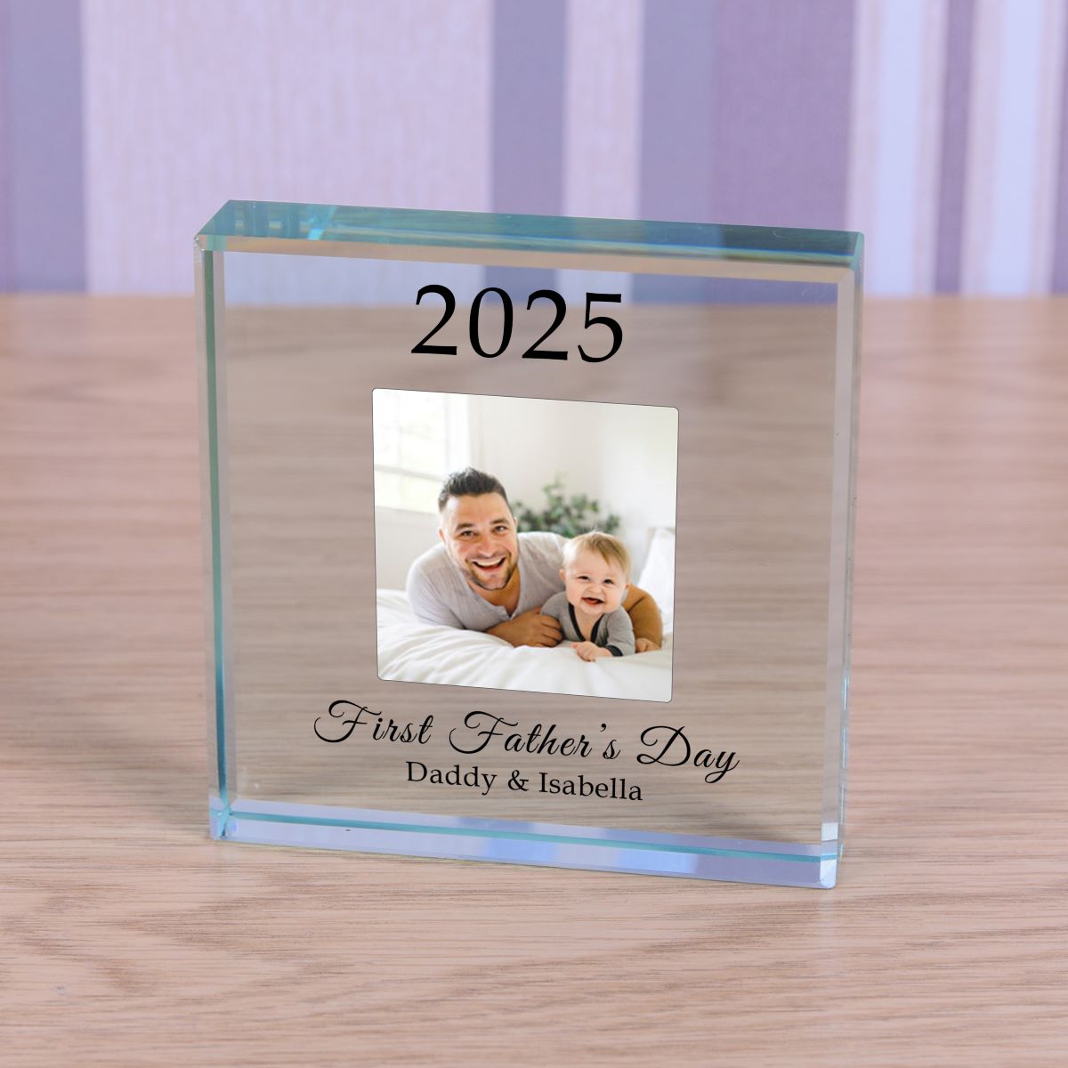 A personalised clear glass square block on a light wood tabletop, featuring engraved text '2025', a custom photo of a father and child, and 'First Father's Day Daddy & Isabella'.