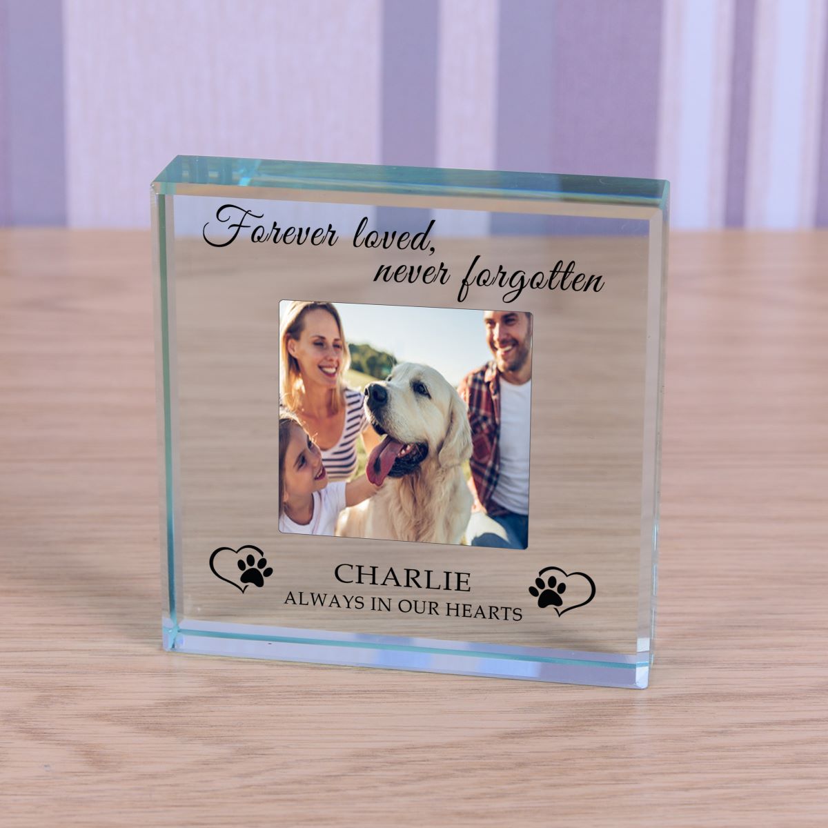 A personalised clear glass keepsake photo block on a light wood tabletop. The block features a custom photo of a man, woman, and their golden retriever dog, with engraved text reading 'Forever loved, never forgotten', 'CHARLIE', and 'ALWAYS IN OUR HEARTS' along with black paw print designs.