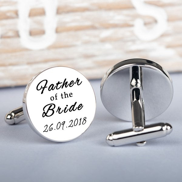 A pair of round silver-toned cufflinks on a light grey surface. One cufflink is shown from the front with black script text reading 'Father of the Bride' and a date.
