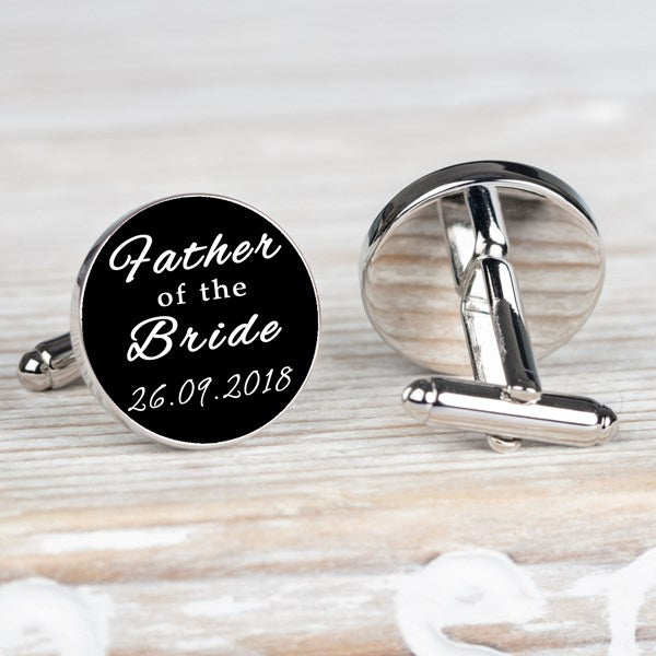 A pair of round silver-toned cufflinks on a light wooden surface. One cufflink is shown from the front with white script text reading 'Father of the Bride' and a date on a black background.