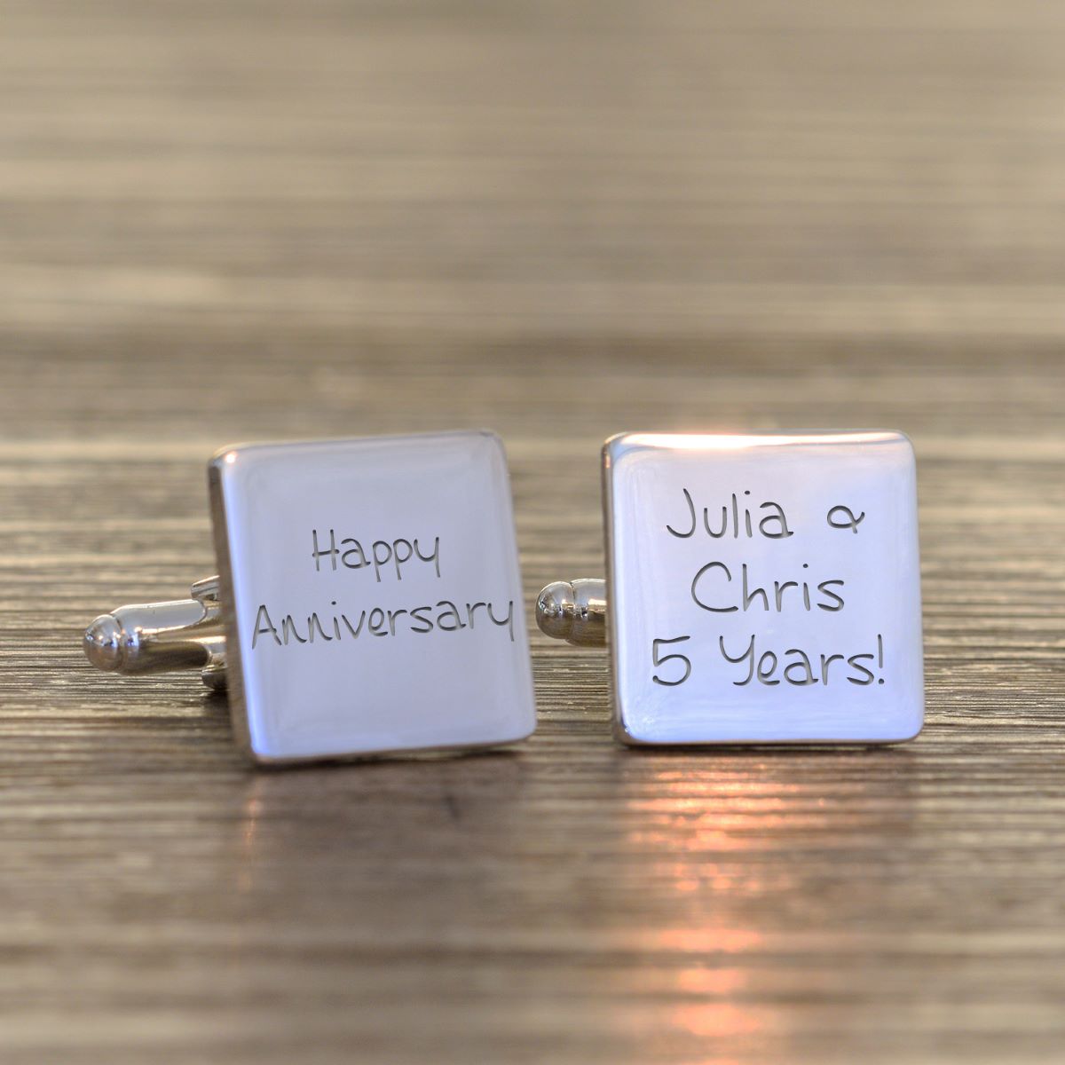 A pair of square silver-toned cufflinks with engraved personalised anniversary messages, displayed on a wooden surface. One cufflink is engraved with 'Happy Anniversary' and the other with 'Julia & Chris 5 Years!'.