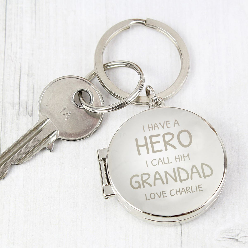 A silver metal keychain with a round engraved tag that reads 'I HAVE A HERO I CALL HIM GRANDAD LOVE CHARLIE,' shown with a key on a light whitewashed wood background.