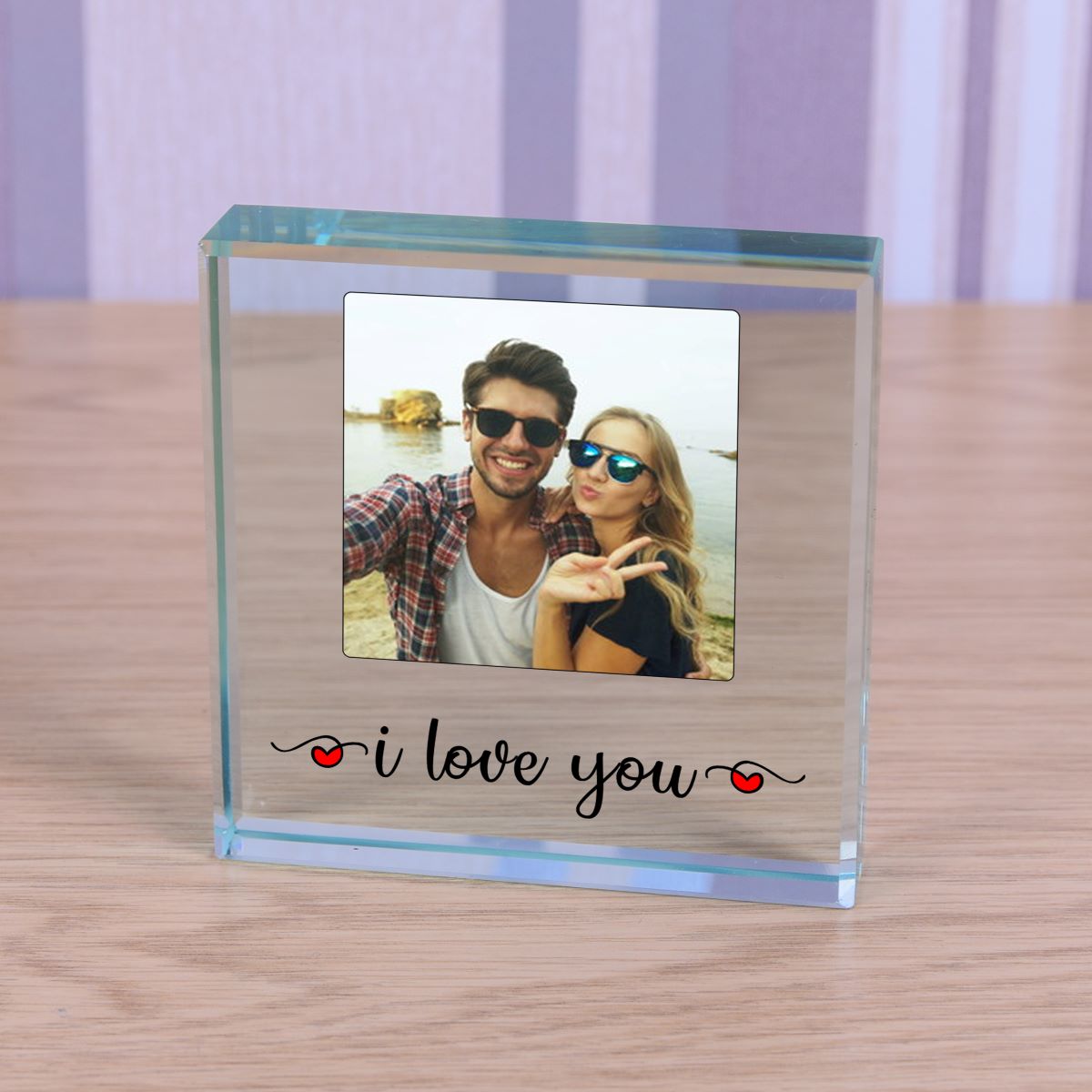 A square clear glass photo block engraved with 'i love you' and small red hearts, with a custom photo of a couple, displayed on a wooden table with a soft purple background.