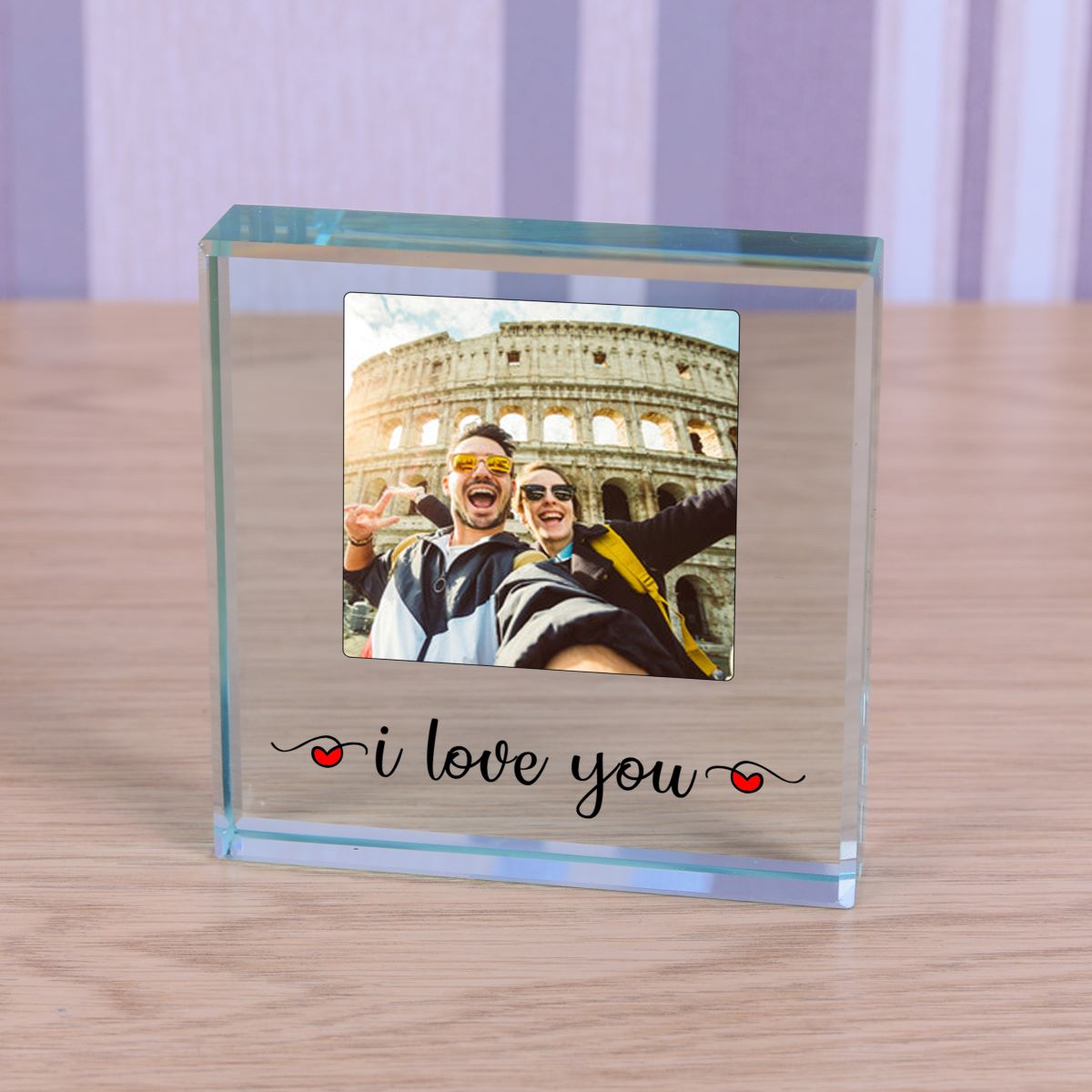 A clear glass photo block on a light wood tabletop, featuring a customisable square photo of a couple in front of the Colosseum and the printed text 'i love you' with small red hearts, set against a soft purple and white striped background.