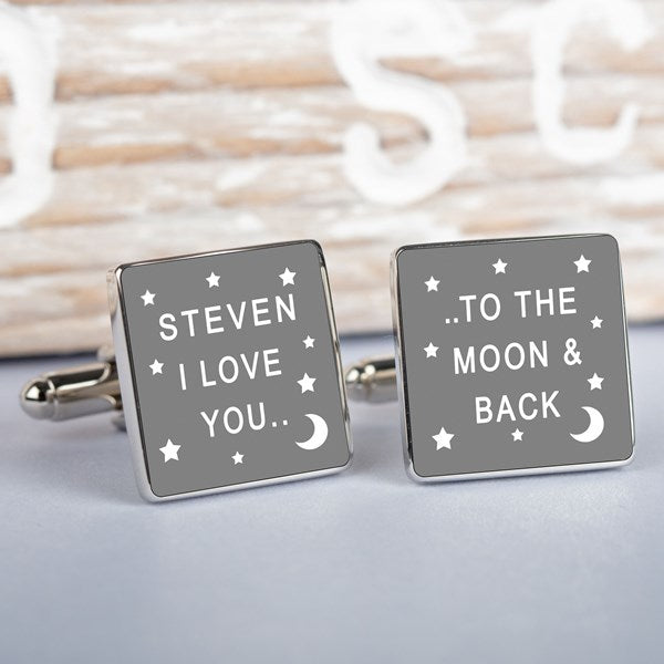 A pair of square silver cufflinks with a grey enamel face, one featuring white text that reads 'STEVEN I LOVE YOU' and the other 'TO THE MOON AND BACK', displayed on a light wooden surface.