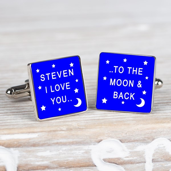 A pair of square silver cufflinks with a bright blue enamel face, one featuring white text that reads 'STEVEN I LOVE YOU' and the other 'TO THE MOON AND BACK', displayed on a light wooden surface.