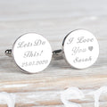 A pair of round silver cufflinks engraved with personalised text, displayed on a light wood surface. One cufflink reads 'Lets Do This! 25.07.25 and the other 'I Love You, Sarah'