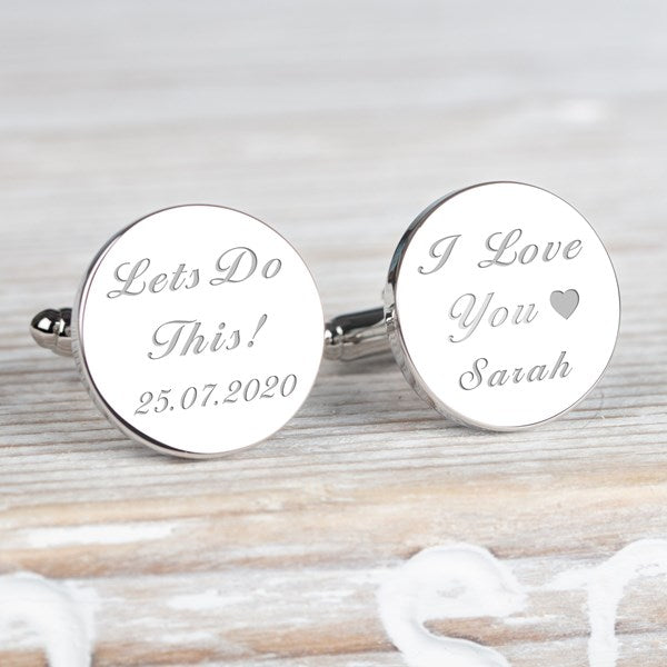 A pair of round silver cufflinks engraved with personalised text, displayed on a light wood surface. One cufflink reads 'Lets Do This! 25.07.25 and the other 'I Love You, Sarah'