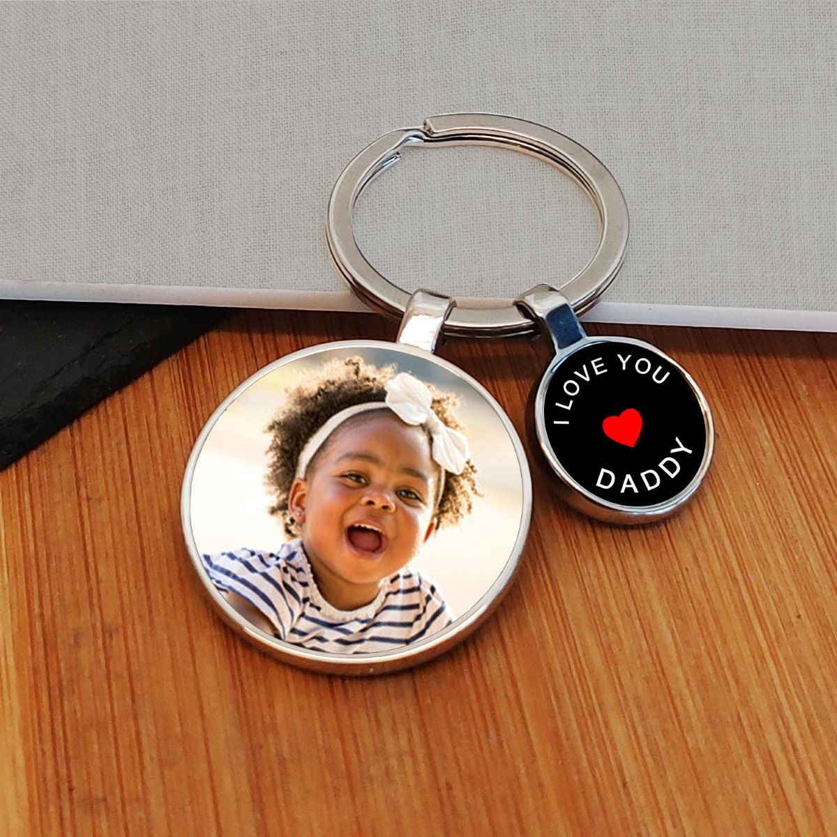 A custom photo keyring with a round metal ring, featuring two charms: one displaying a photo of a little girl, and the other showing a black background with white writing saying 'I LOVE YOU DADDY' and a red heart. The keychain is displayed on a wooden surface.