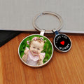 A custom photo keyring with a round metal ring, featuring two charms: one displaying a photo of a little girl, and the other showing a black background with white writing saying 'I LOVE YOU MUMMY' and a red heart. The keychain is displayed on a wooden surface.