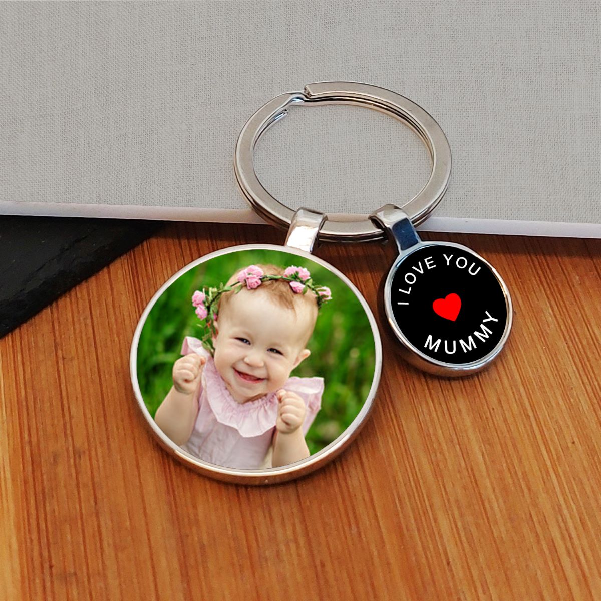 A custom photo keyring with a round metal ring, featuring two charms: one displaying a photo of a little girl, and the other showing a black background with white writing saying 'I LOVE YOU MUMMY' and a red heart. The keychain is displayed on a wooden surface.