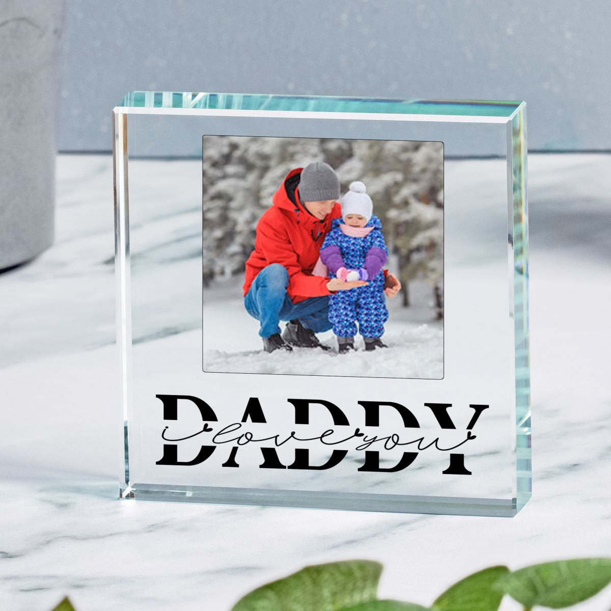 A personalised clear glass block plaque display on a light marble surface, featuring a photo of a man and a child, and engraved text reading 'Daddy I love you'.
