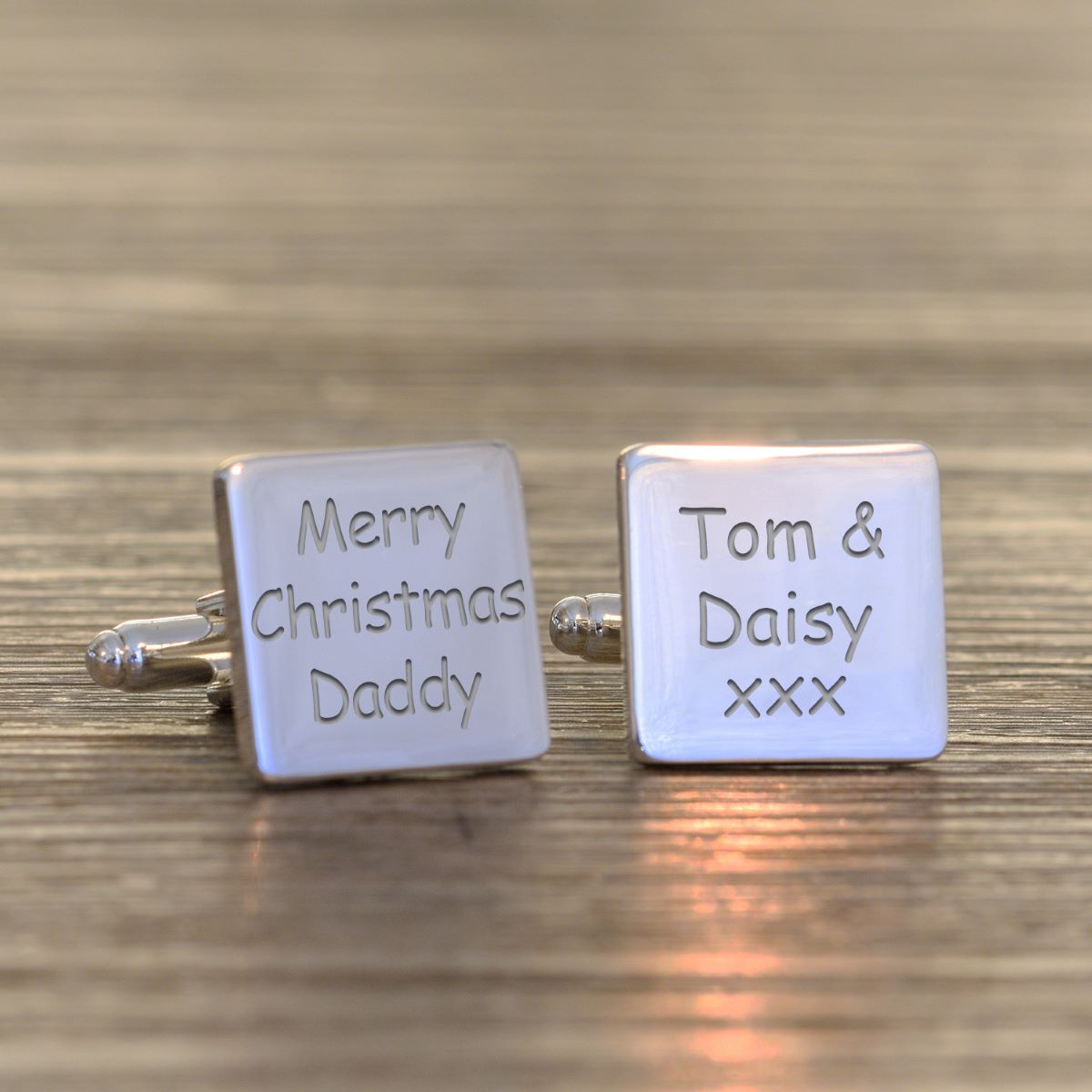 A pair of square silver cufflinks resting on a wooden surface. One cufflink is engraved with 'Merry Christmas Daddy' and the other with 'Tom & Daisy xxx'.