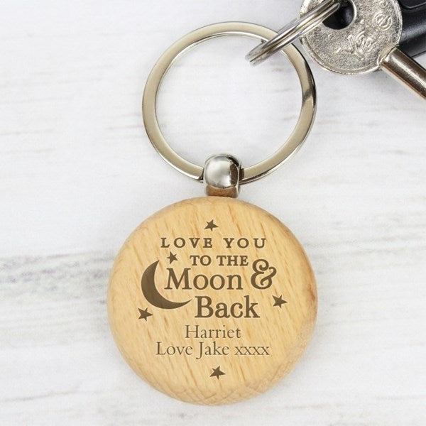 A personalized round wooden keychain with engraved text reading 'LOVE YOU TO THE Moon & Back Harriet Love Jake xxxx' and small moon and star designs, attached to a silver metal keyring with keys on a light wood surface.