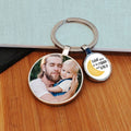 A round metal keyring with a custom photo of man and a young child on one charm and a yellow crescent moon with the text 'LOVE you to the MOON and BACK' on the other, displayed on a wooden surface.