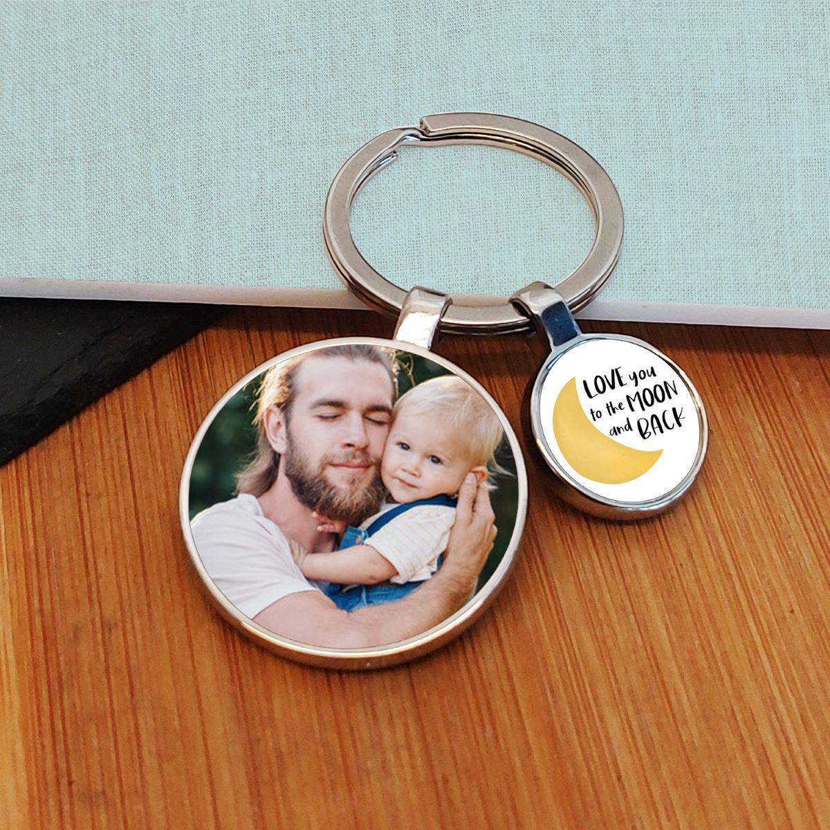 A round metal keyring with a custom photo of man and a young child on one charm and a yellow crescent moon with the text 'LOVE you to the MOON and BACK' on the other, displayed on a wooden surface.