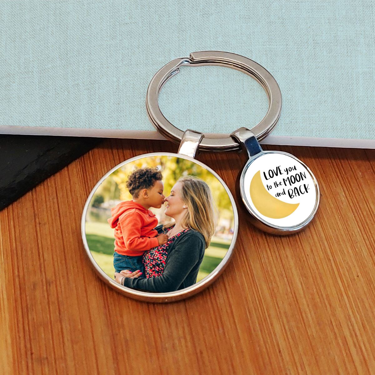 A round metal keyring with a custom photo of a lady and child on one charm and a yellow crescent moon with the text 'LOVE you to the MOON and BACK' on the other, displayed on a wooden surface.