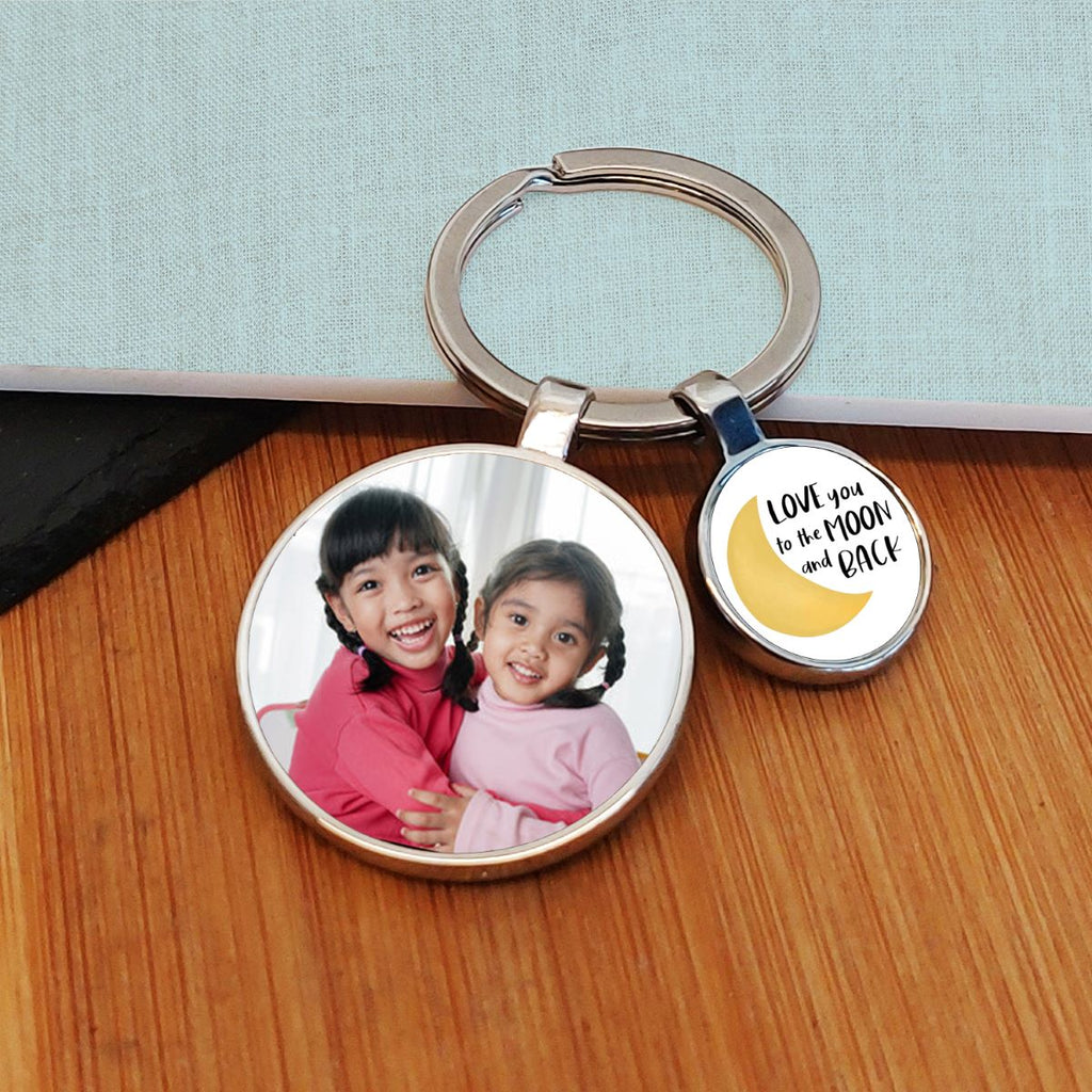 A round metal keyring with a custom photo of two young girls on one charm and a yellow crescent moon with the text 'LOVE you to the MOON and BACK' on the other, displayed on a wooden surface.