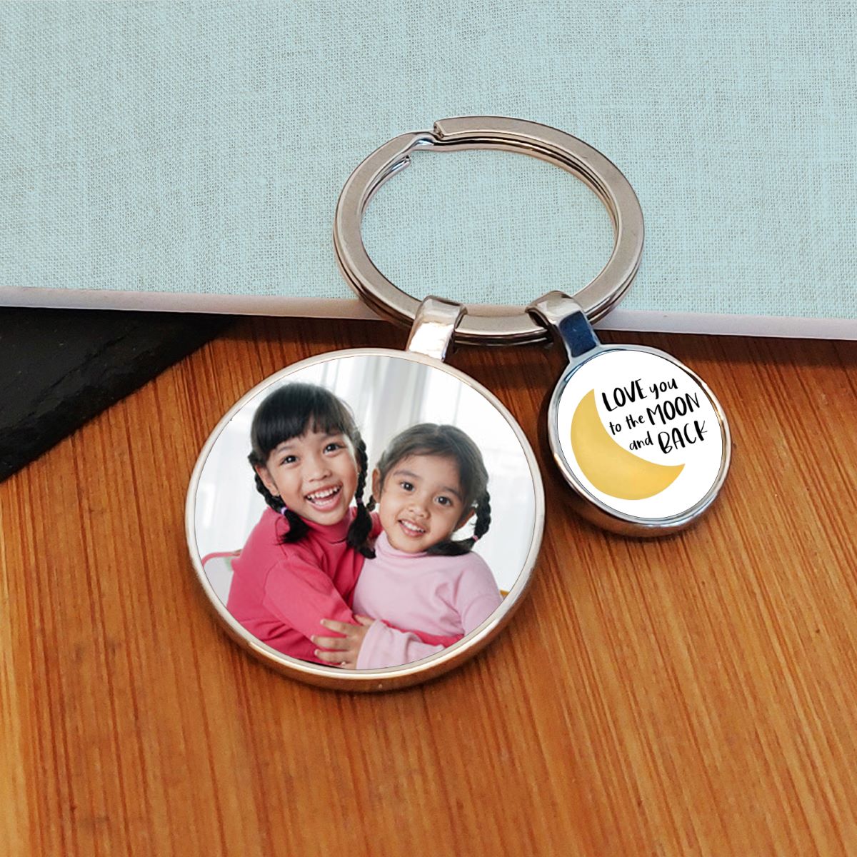 A round metal keyring with a custom photo of two young girls on one charm and a yellow crescent moon with the text 'LOVE you to the MOON and BACK' on the other, displayed on a wooden surface.
