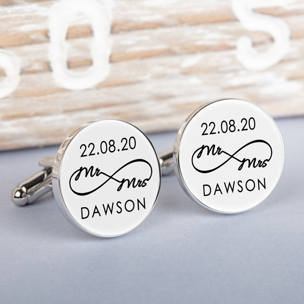 A pair of round silver-toned cufflinks on a light grey surface. Both cufflink faces are white with a black infinity symbol with small wording 'Mr Mrs' along with a date and surname