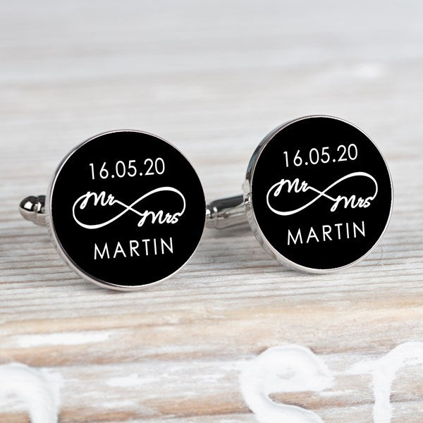 A pair of round silver-toned cufflinks on a light grey surface. Both cufflink faces are black with a white infinity symbol with small wording 'Mr Mrs' along with a date and surname