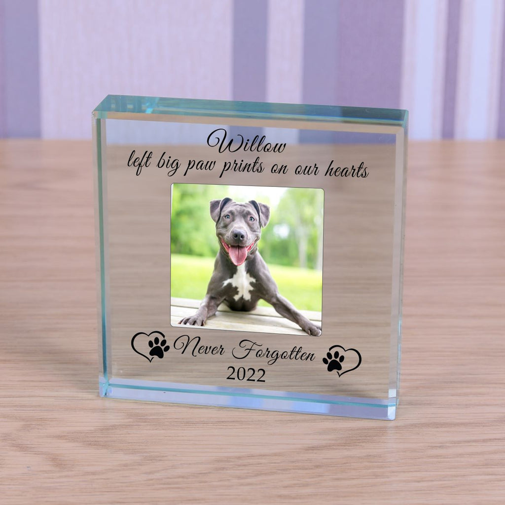 A square personalised memorable clear glass block display on a light wood surface, featuring a photo of a dog and engraved text reading 'Willow left big paw prints on our hearts, never forgotten 2022' with paw prints and love hearts on either side.
