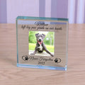 A square personalised memorable clear glass block display on a light wood surface, featuring a photo of a dog and engraved text reading 'Willow left big paw prints on our hearts, never forgotten 2022' with paw prints and love hearts on either side.
