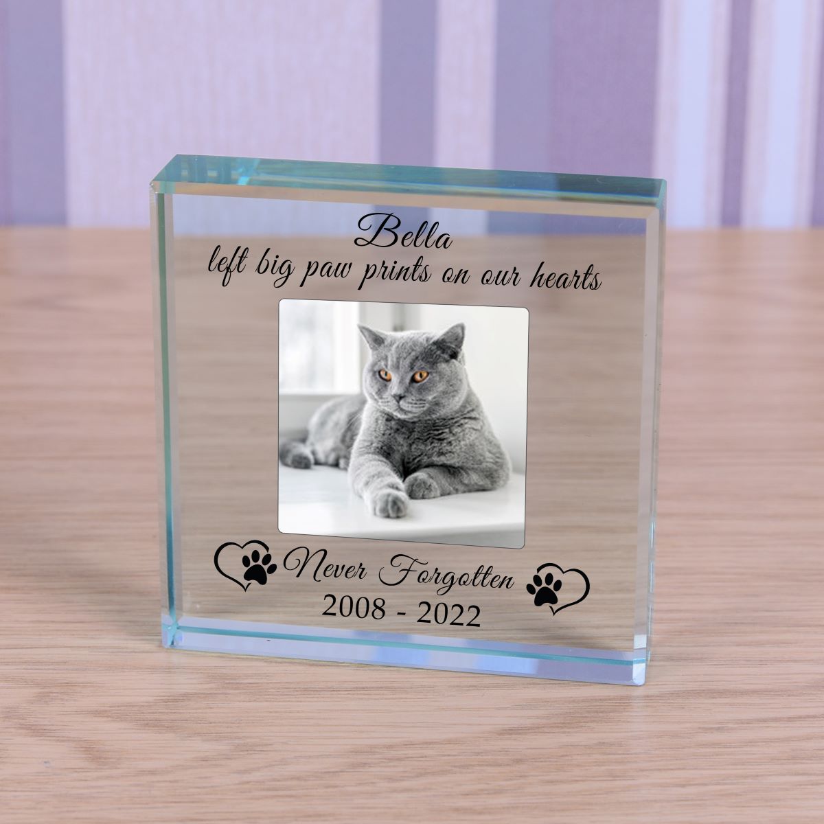A square personalised memorable clear glass block display on a light wood surface, featuring a photo of a cat and engraved text reading 'Bella left big paw prints on our hearts, never forgotten 2008-2022' with paw prints and love hearts on either side.
