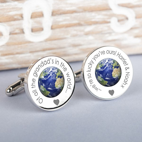 A pair of round silver cufflinks featuring a detailed globe of Earth in the center, surrounded by engraved personalised text 'Of all the grandad's in the world..' on one and '...we're so lucky you're ours!  Harriet and Noah x' on the other and a small heart, displayed on a neutral grey surface.