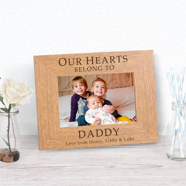 A personalised wooden photo frame displayed on a light wood surface, featuring a photo three children and engraved text reading 'OUR HEARTS BELONG TO DADDY, Love from Henry, Lily & Luke'.
