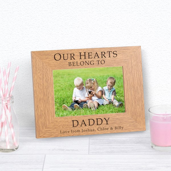 A personalised wooden photo frame displayed on a light wood surface, featuring a photo three children and engraved text reading 'OUR HEARTS BELONG TO DADDY, Love from Joshua, Chloe & Billy'.
