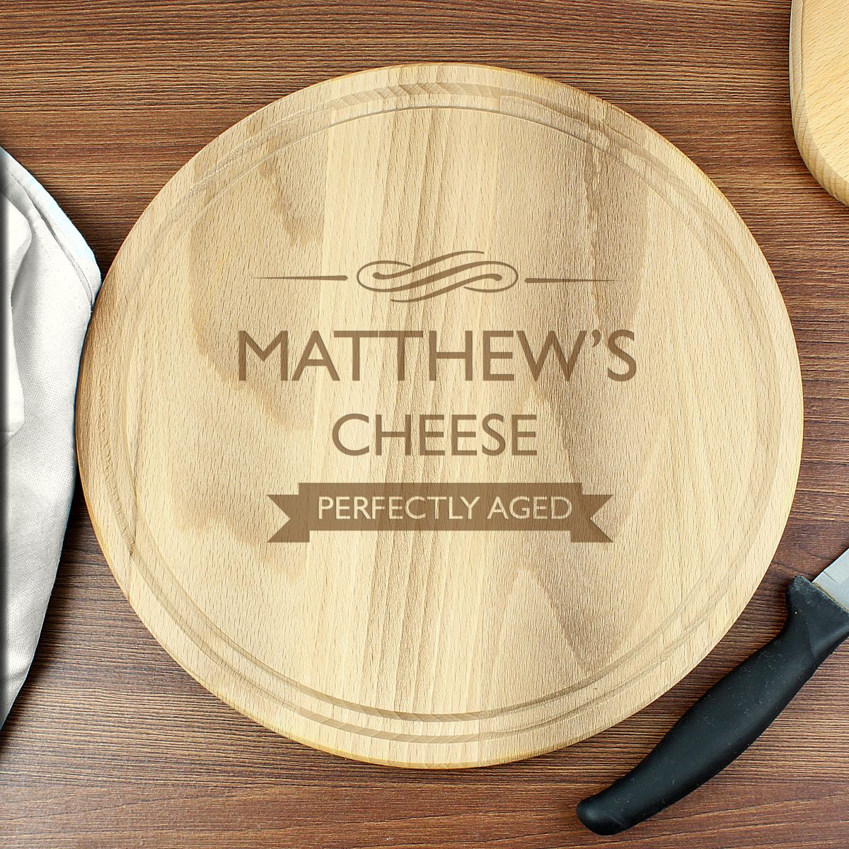 A round wooden cheese board with engraved text reading 'MATTHEW'S CHEESE' and 'PERFECTLY AGED' on a wooden tabletop, with a knife and cloth partially visible around it.