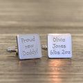 A pair of square silver cufflinks on a wooden surface. One cufflink is engraved with 'Proud new Daddy!' and the other with 'Olivia Jones 6lbs 2oz'.
