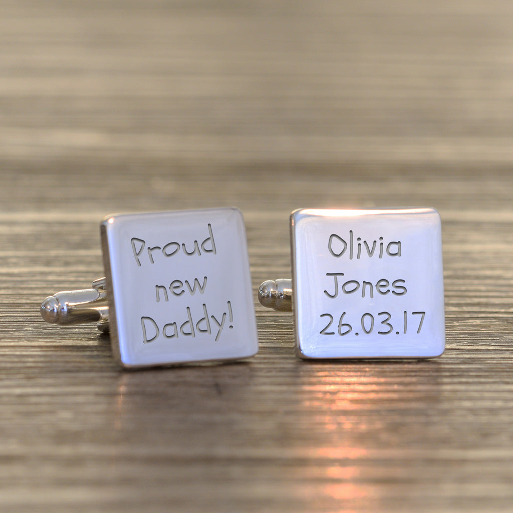 A pair of square silver cufflinks on a wooden surface. One cufflink is engraved with 'Proud new Daddy!' and the other with 'Olivia Jones 26.03.17'.