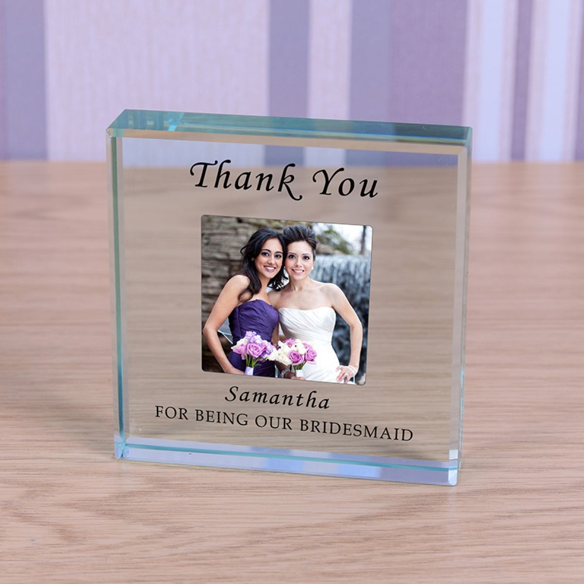 A personalized clear glass keepsake photo block on a light wood tabletop. The block is engraved with 'Thank You' at the top and 'Samantha FOR BEING OUR BRIDESMAID' at the bottom, featuring a custom photo of a bride and bridesmaid.