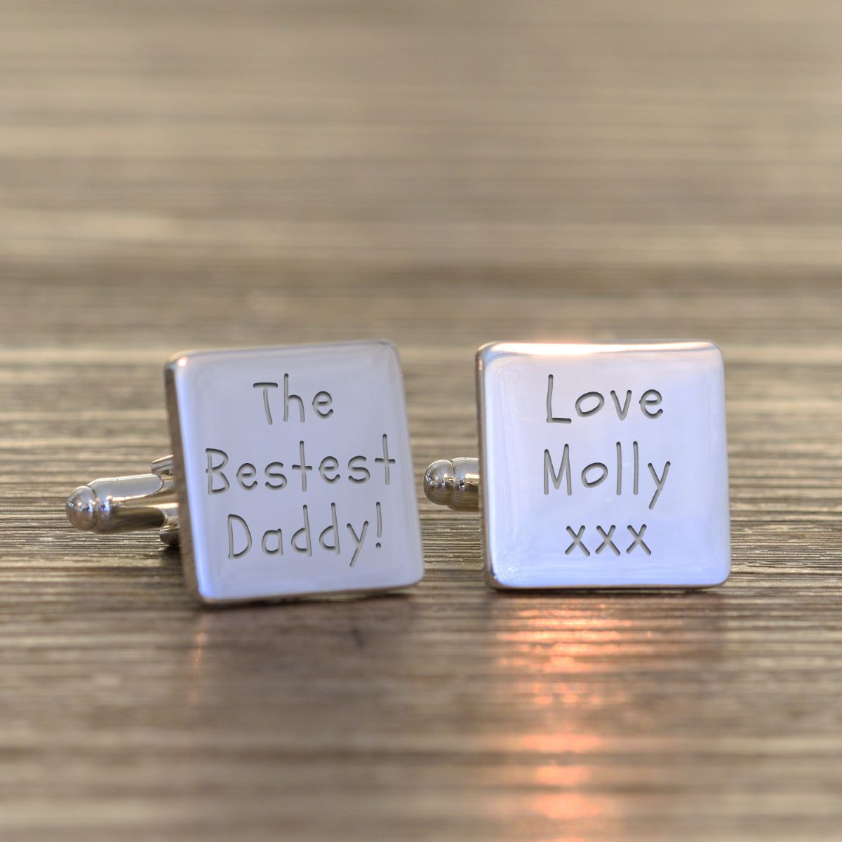 A pair of square silver cufflinks engraved with personalised messages, one reading 'The Bestest Daddy!' and the other 'Love Molly xxx', displayed on a wooden surface.