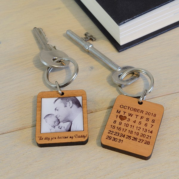 Two engraved wooden keyrings with metal keys on a light wood tabletop. One keyring shows a calendar for October 2018 with a heart over the number 2, and the other says has an image of a man holding a baby with the words 'The day you became my Daddy!
