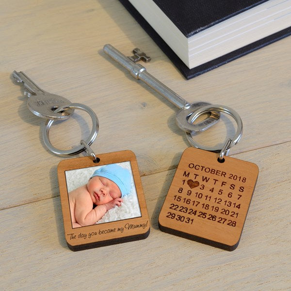 Two engraved wooden keyrings with metal keys on a light wood tabletop. One keyring shows a calendar for October 2018 with a heart over the number 2, and the other says has an image of a baby with the words 'The day you became my Mummy!