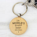 A personalized wooden keychain with a round light tan tag engraved with 'The WORLD'S Greatest DADDY MICHAEL' in dark lettering, attached to a silver metal split keyring with keys on a white wood background.