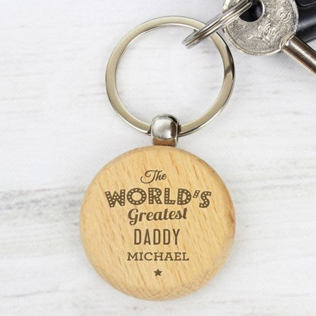 A personalized wooden keychain with a round light tan tag engraved with 'The WORLD'S Greatest DADDY MICHAEL' in dark lettering, attached to a silver metal split keyring with keys on a white wood background.