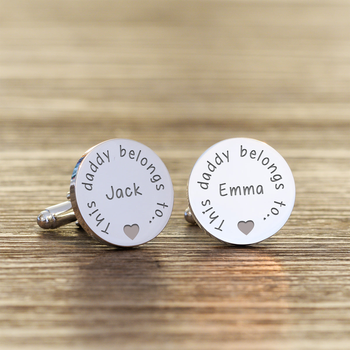 Silver cufflinks with the engraved text 'This daddy belongs to.. Jack' on one and 'This daddy belongs to.. Emma' on the other with a heart silhouette displayed on a wooden surface