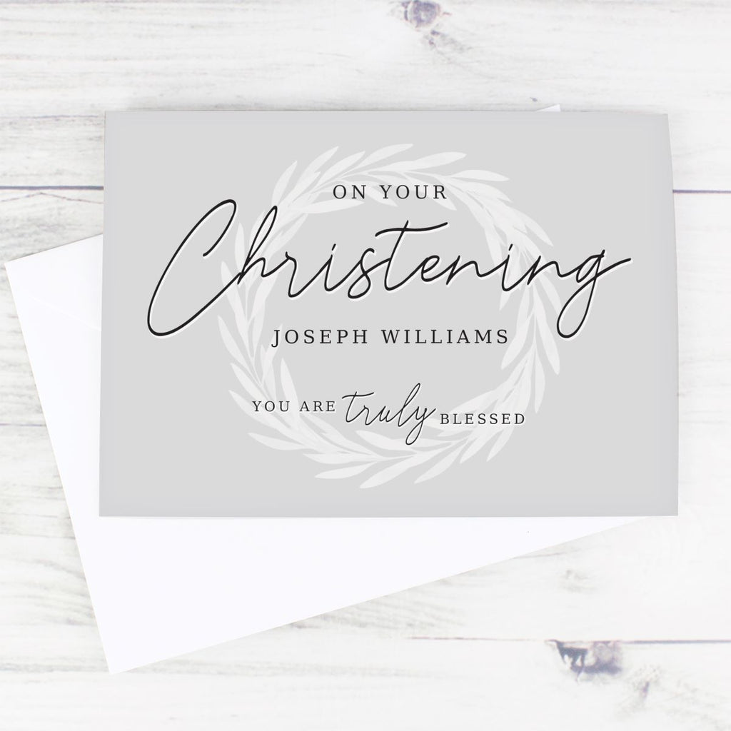A personalized Christening greeting card with a white envelope, displayed on a light whitewashed wooden surface. The card is light gray with elegant black script reading 'On Your Christening Joseph Williams You Are Truly Blessed' surrounded by a delicate white leaf wreath design.