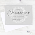 A personalized Christening greeting card with a white envelope, displayed on a light whitewashed wooden surface. The card is light gray with elegant black script reading 'On Your Christening Joseph Williams You Are Truly Blessed' surrounded by a delicate white leaf wreath design.