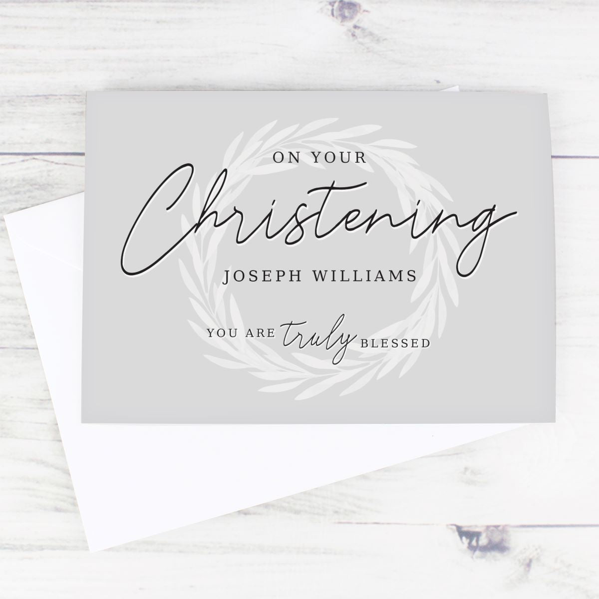 A personalized Christening greeting card with a white envelope, displayed on a light whitewashed wooden surface. The card is light gray with elegant black script reading 'On Your Christening Joseph Williams You Are Truly Blessed' surrounded by a delicate white leaf wreath design.