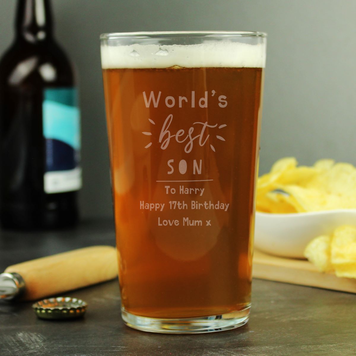 Pint glass with engraved message, beer bottle, and snacks on a wooden surface