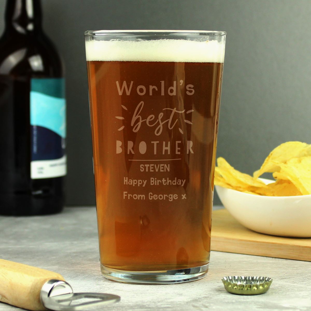 Personalized beer glass with 'World's best brother' text, filled with beer, on a table with a bottle and chips in the background.